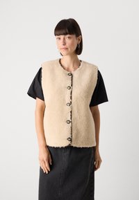 Beige Fleeceweste mit Rundhalsauschnitt, fünf Knopfverschluss und kontrastierenden Knopfdetails. Getragen über einem schwarzen T-Shirt mit kurzen Ärmeln.