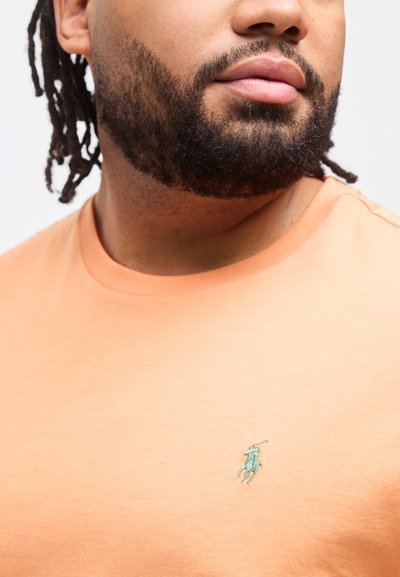 Άνδρας με γενειάδα και dreadlocks, που φοράει ροδακινί μπλουζάκι με στρογγυλή λαιμόκοψη και μικρό πράσινο λογότυπο παίκτη πόλο στο στήθος.