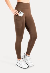 Bruine leggings met hoge taille en een zijzak, gemaakt van rekbare stof. Voorzien van contrasterende stiksels en een gladde, gestructureerde afwerking.