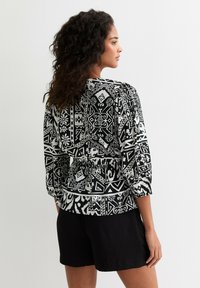 Blouse à motifs noir et blanc avec des manches trois-quarts, taille élastique et motifs géométriques complexes. Tissu lisse avec une coupe décontractée.