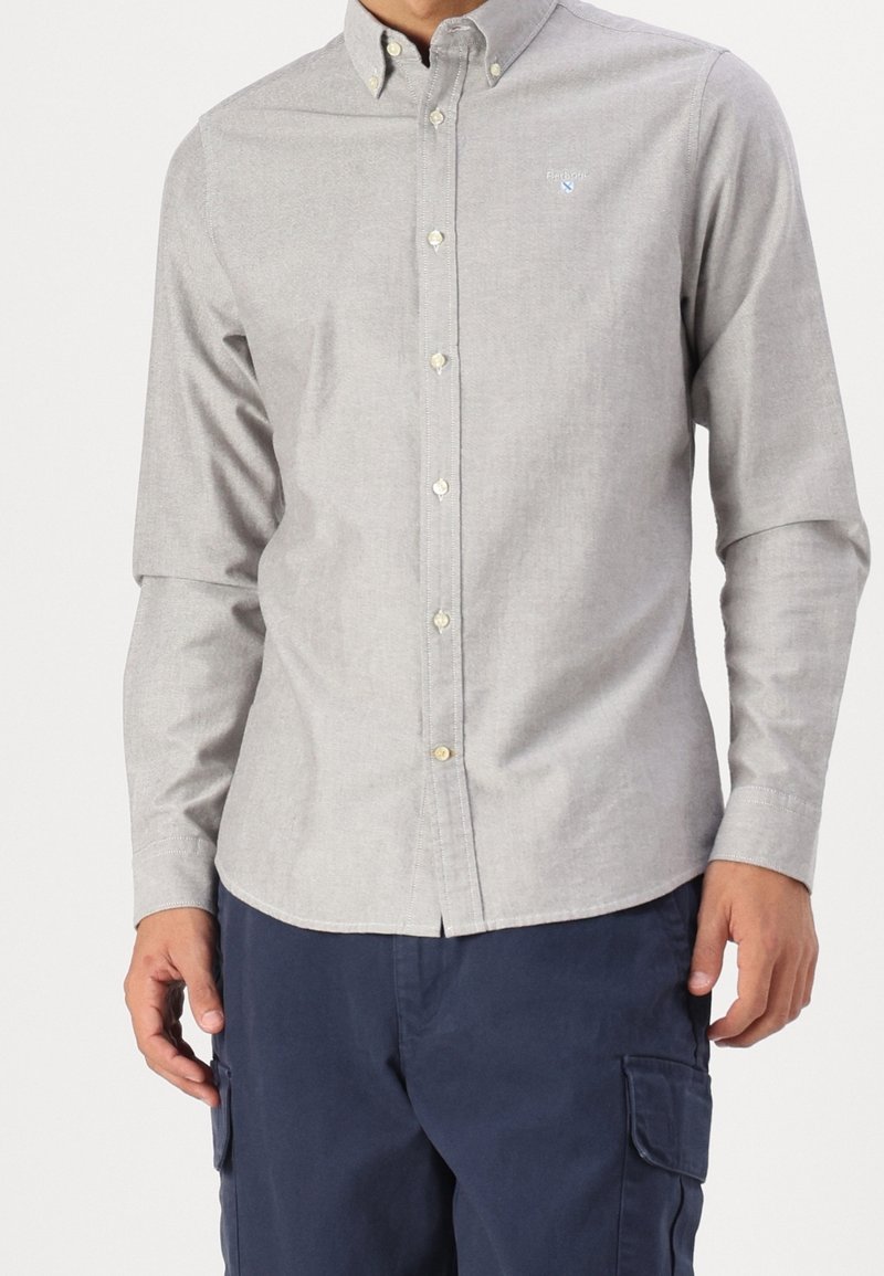Homme portant une chemise gris clair à manches longues boutonnée et un pantalon cargo bleu foncé, debout les mains détendues le long du corps.