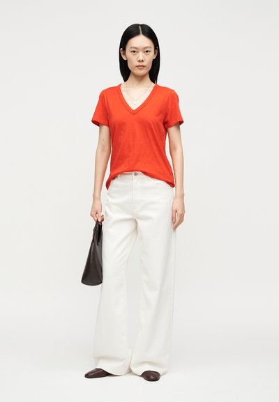 Γυναίκα με κόκκινο μπλουζάκι V-λαιμόκοψη, φαρδιά λευκά παντελόνια, καφέ παπούτσια, κρατώντας μαύρη τσάντα σε απλό φωτεινό φόντο.