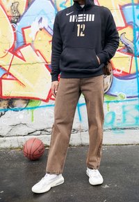 Persona con felpa nera e pantaloni marroni in piedi sul marciapiede con un pallone da basket vicino a un muro graffitato colorato.