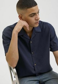 Camisa de manga corta azul marino en tejido texturizado con cuello de punta, dos botones en la parte delantera y un corte relajado. Combinada con jeans de mezclilla.