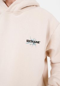Beige Hoodie mit einer vorderen Kängurutasche, versehen mit einem schwarzen "SIXTH JUNE" Logo und einem blauen Blitzdesign, das auf der Brust gestickt ist.