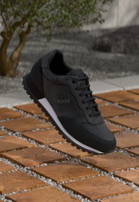 Zwarte sportieve sneaker met een bovenwerk van mesh, platte veters en een witte tussenzool, voorzien van een gestructureerde zool voor grip en een logo aan de zijkant.
