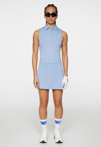 Abito da golf azzurro chiaro con colletto, design senza maniche, vita aderente e gonna svasata. Accompagnato da guanti bianchi e occhiali da sole sportivi.