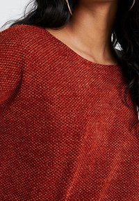Pull rouge tricoté avec un motif texturé et un col large, présentant une coupe ample et un matériau doux. Accents de nuances de rouge foncé et clair.