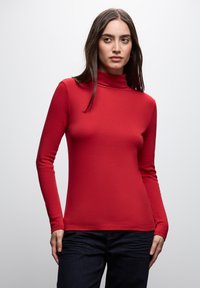 Femme aux longs cheveux foncés portant un pull à col roulé rouge et un jean foncé, sur un fond clair uni.