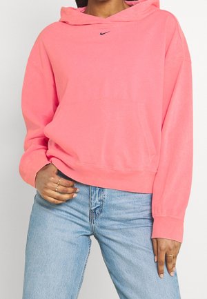 Femme portant un sweat à capuche rose ample Nike et un jean bleu clair avec des bagues aux doigts, main reposant sur la hanche et la cuisse.