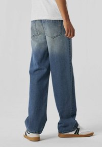 Jeans de mezclilla azul con corte holgado, lavado deslavado, diseño de cinco bolsillos y detalles cosidos en los bolsillos traseros. Llevados con zapatillas blancas.