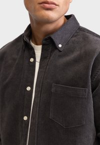 Dunkelgraues Cordhemd mit Button-Down-Kragen, Brusttasche und strukturiertem Rippmuster. Besitzt helle Knöpfe an der Knopfleiste.