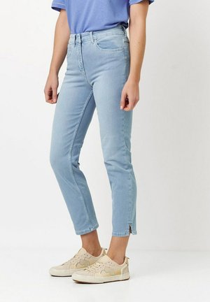 Hellblaue Jeans mit geradem Bein, featuring seitliche Schlitzdetail am Saum, kombiniert mit beigen Sneakers und einem blauen Shirt.