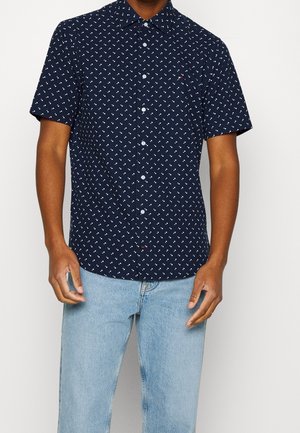 Homme portant une chemise bleu marine à manches courtes avec un petit motif géométrique blanc et un jean bleu clair.