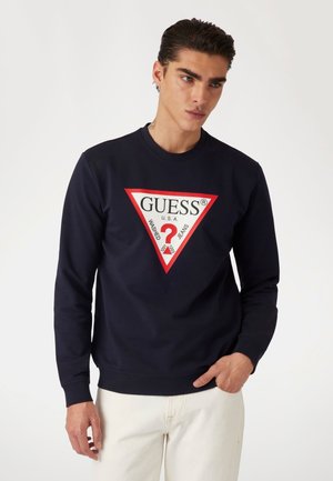 Sudadera azul marino con un logotipo triangular rojo que presenta "GUESS® USA" y "Washed Jeans" en texto blanco y negro. Mangas largas, cuello redondo.