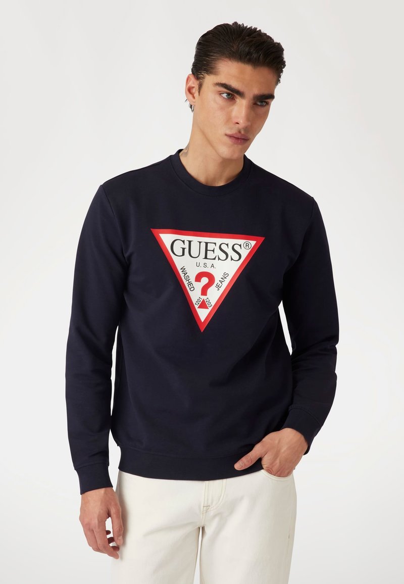 Felpa blu navy con un logo triangolare rosso che presenta "GUESS® USA" e "Washed Jeans" in testo bianco e nero. Maniche lunghe, colletto a girocollo.