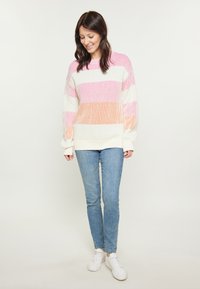 Gebreide trui met horizontale strepen in roze, oranje en wit. Heeft een relaxte pasvorm en geribde manchetten, in combinatie met lichtblauwe jeans en witte schoenen.