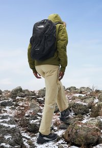 Mochila negra con logo, chaqueta verde oliva, pantalones beiges y zapatos negros. Terreno rocoso con parches de nieve bajo un cielo azul claro.