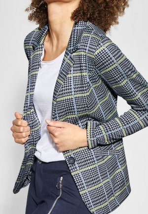 Frau trägt ein weißes Oberteil, karierten Blazer mit gelben Linien und marineblaue Hosen mit einem großen silbernen Reißverschlussdetail an der Seite.