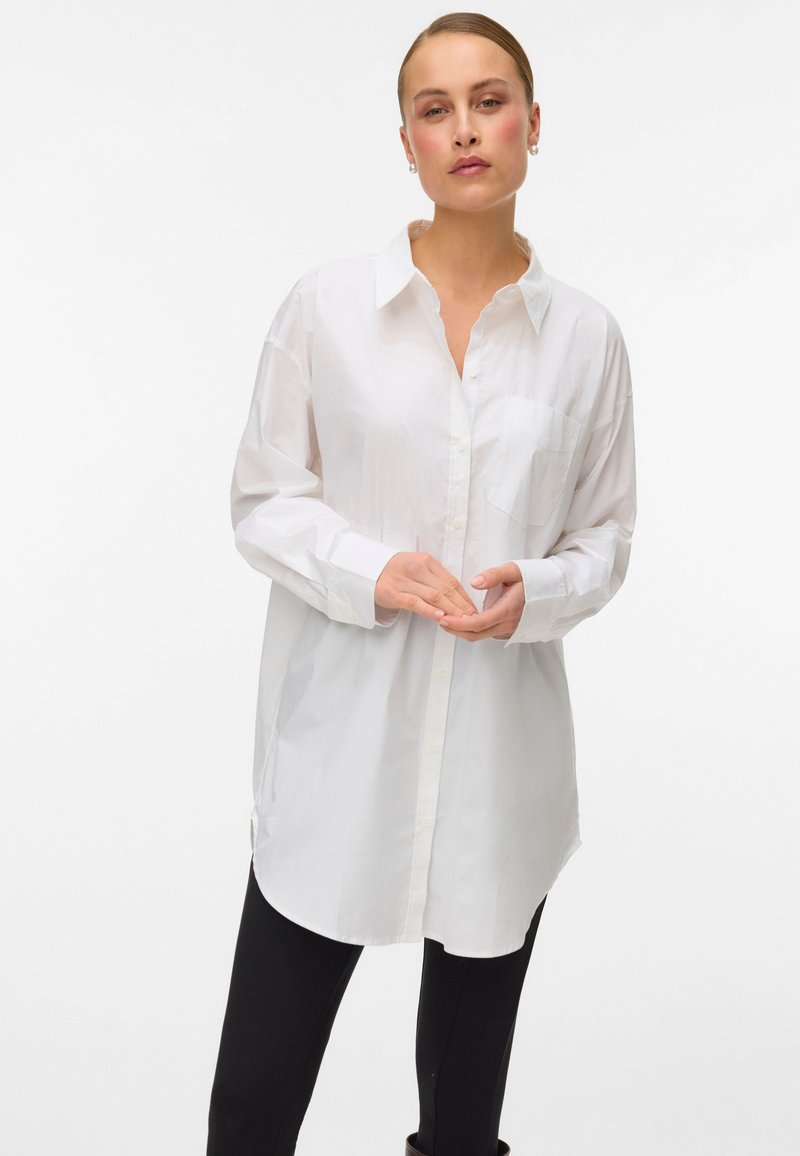 Hvit langermet skjorte med krage, en brystlomme og splitt i sidene; brukt over svarte leggings. Glatt stofftekstur.