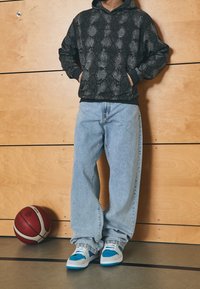 Felpa nera con un motivo a serpente testurizzato, jeans chiari larghi e scarpe con accenti blu. Un pallone da basket è posato vicino ai piedi.