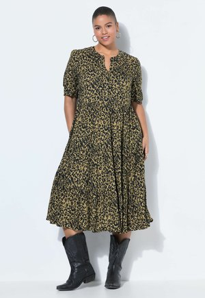 Kobieta pozująca w sukience z nadrukiem w cętki o długości do połowy łydki, z półdługimi rękawami i czarnych kowbojskich butach na białym tle.