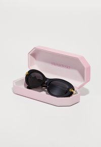 Melni saulesbrilles ar ovālām rāmēm, rotātas ar zelta akcentiem, rozā astoņstūrīgā gadījumā ar uzrakstu "Swarovski".