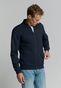Marineblauwe gezipte jas met een textuurpatroon, staande kraag en zijzakken. Draag het over een wit shirt, gecombineerd met lichtblauwe jeans.