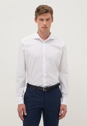 Homme portant une chemise blanche ajustée, un pantalon bleu marine et une ceinture noire, debout devant un fond clair uni.