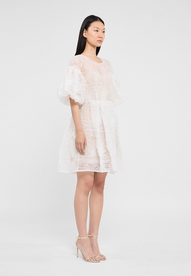Robe blanche texturée avec des manches ballon, présentant une coupe ample et un design superposé ; accompagnée de sandales à talons hauts et à lanières.