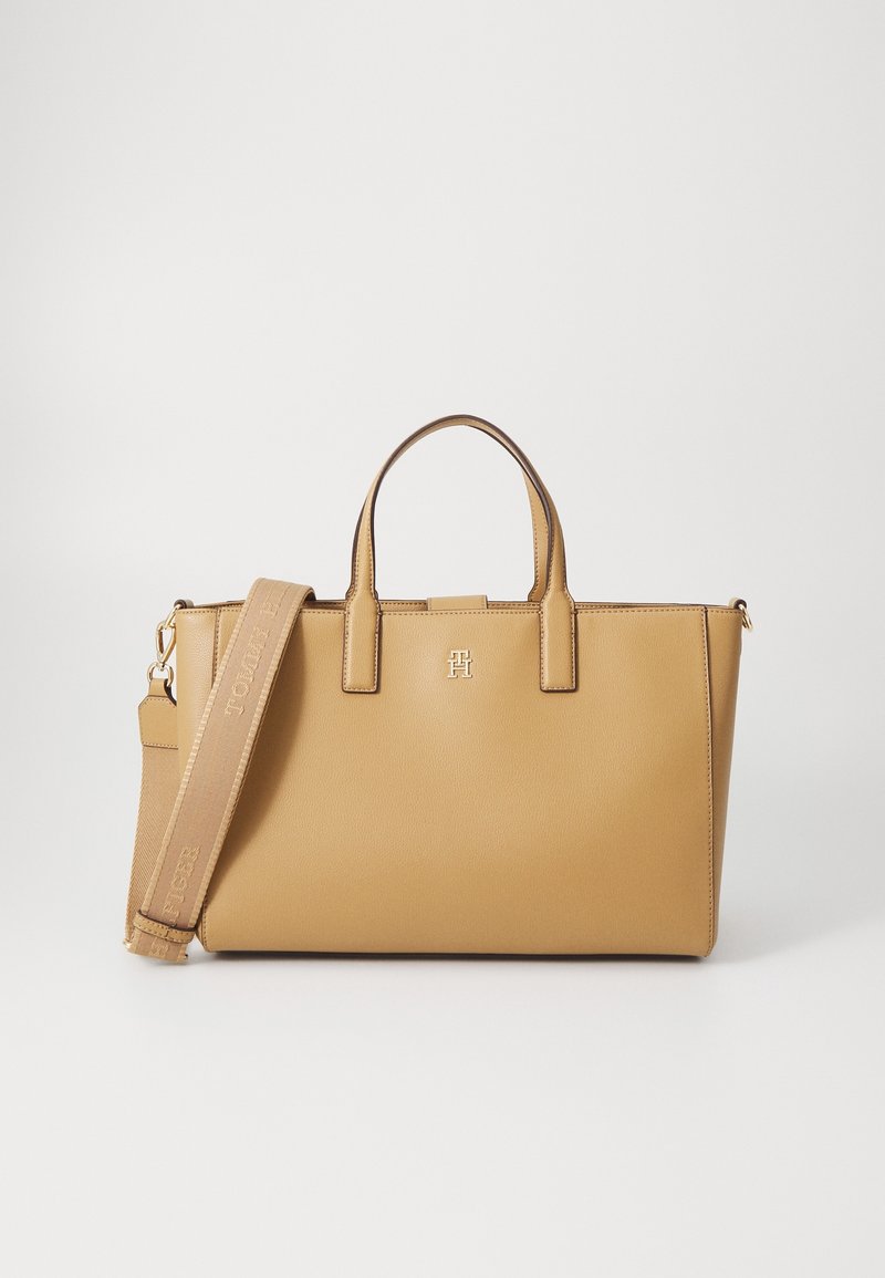 Sac fourre-tout en cuir couleur tabac avec deux poignées supérieures et une bandoulière large amovible embossée du nom de la marque sur fond blanc.