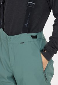 Vattentåliga skidbyxor i teal med justerbara svarta hängslen, fickor med dragkedja, slät textur och knappstängning.