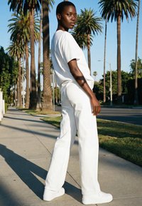 Persoon in een wit T-shirt, broek en sneakers die op een trottoir staat dat is omzoomd met hoge palmbomen in fel daglicht.