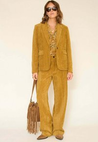 Femme portant un costume en velours côtelé moutarde avec une chemise à motifs, un sac à main marron à franges, des lunettes de soleil et des chaussures marron pointues, debout devant un fond beige.