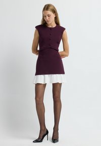 Robe ajustée avec un haut violet comportant cinq boutons et des épaulettes structurées, superposée à une jupe blanche évasée ; associée à des talons noirs.