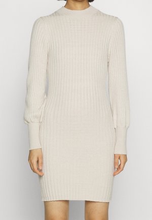 Vrouw draagt een nauwsluitende, lange mouw, crèmekleurige gebreide jurk met geribbelde structuur, met omgeslagen mouwen en een ronde hals.