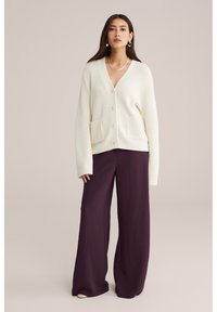 Donna con lunghi capelli scuri che indossa un cardigan a coste color crema con bottoni e pantaloni larghi di un viola scuro, in piedi contro uno sfondo neutro.