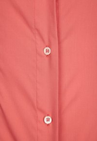 Camisa de botones color coral con una textura suave; presenta botones blancos y costuras en contraste a lo largo de los bordes.