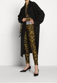 Schwarzer Mantel über einem gemusterten Top und Leggings mit goldenen und schwarzen barocken Motiven, ergänzt durch schwarze spitze Absätze.