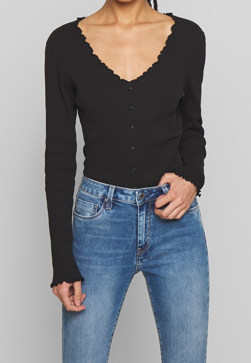 Body en tissu côtelé noir à manches longues avec encolure et poignets festonnés, doté d'une patte de boutonnage sur le devant. Assorti à un jean en denim bleu.