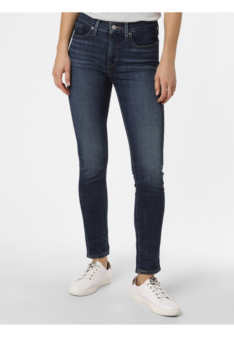 Femme portant un jean skinny bleu foncé et des baskets blanches avec un détail imprimé léopard au talon, sur un fond uni.