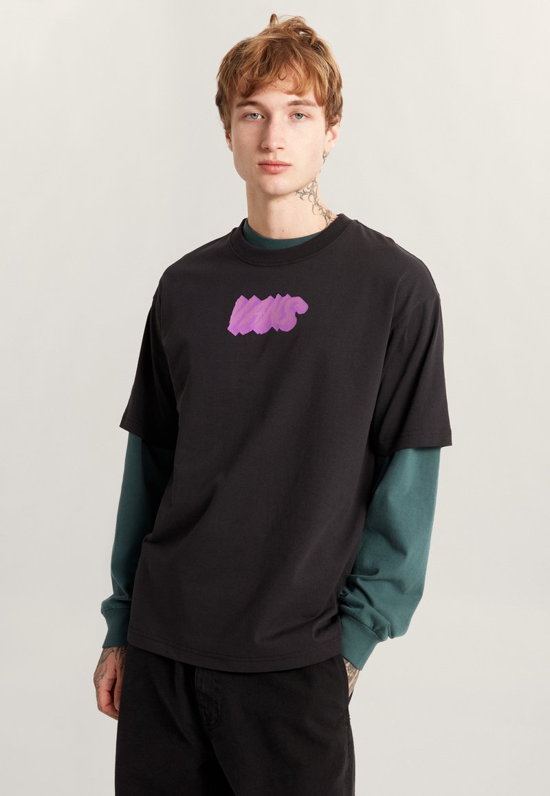 Czarny bawełniany t-shirt z fioletowym logo graficznym z przodu. Nałożony na zieloną koszulę z długim rękawem. Luźny krój, krótkie rękawy.