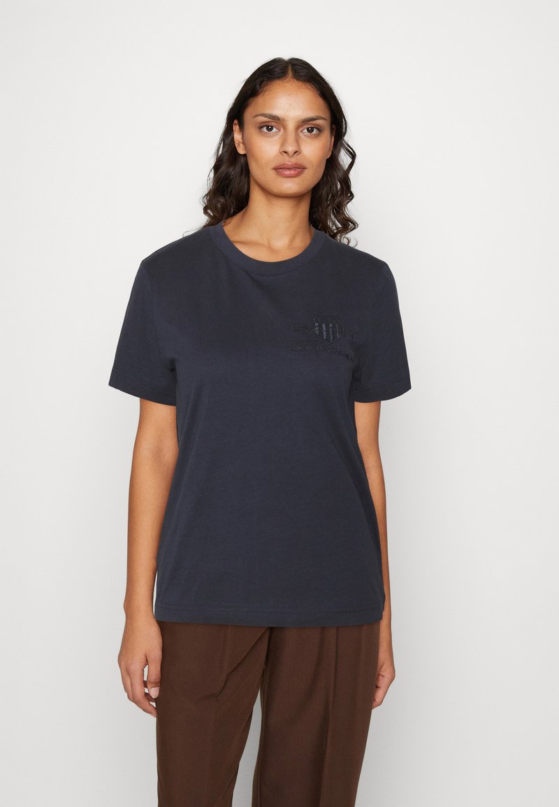Camiseta de algodón azul marino con cuello redondo y mangas cortas, que presenta un logo sutil en relieve en el pecho. Combinada con pantalones marrones.