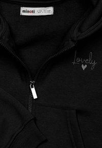 Melnā kapucēta jaka ar rāvējslēdzēju, izgatavota no mīkstas auduma. Ar sudraba rāvējslēdzēju un izšuvumu "Lovely" tekstā ar sirds detaļu uz krūtīm.