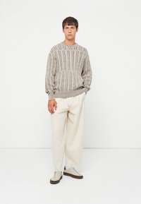 Dicker Strickpullover in Hellgrau mit vertikalen Streifen, geripptem Rundhalsausschnitt und Bündchen. Kombiniert mit locker sitzenden, cremefarbigen Hosen und Turnschuhen.