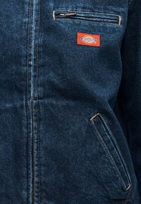 Σκούρο μπλε τζιν μπουφάν με φερμουάρ, κεκλιμένες εμπρόσθιες τσέπες και πορτοκαλί ετικέτα Dickies στην τσέπη στο στήθος.