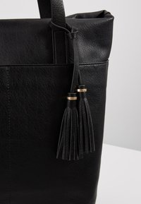 Sac à main en cuir noir avec deux pampilles pendantes aux accents dorés sur un fond clair uni.