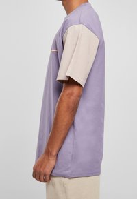 Tričko s krátkým rukávem s levandulovým tělem a béžovými rukávy, s texturovanou tkaninou a malým oranžovým akcentem na hrudi.