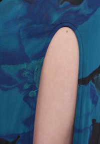 Μπλε ύφασμα με αφηρημένα σχέδια διαθέτει μία ανοίγμα στο ύψος του ώμου. Η υφή φαίνεται ομαλή, αναδεικνύοντας τον μοναδικό σχεδιασμό του ενδύματος.