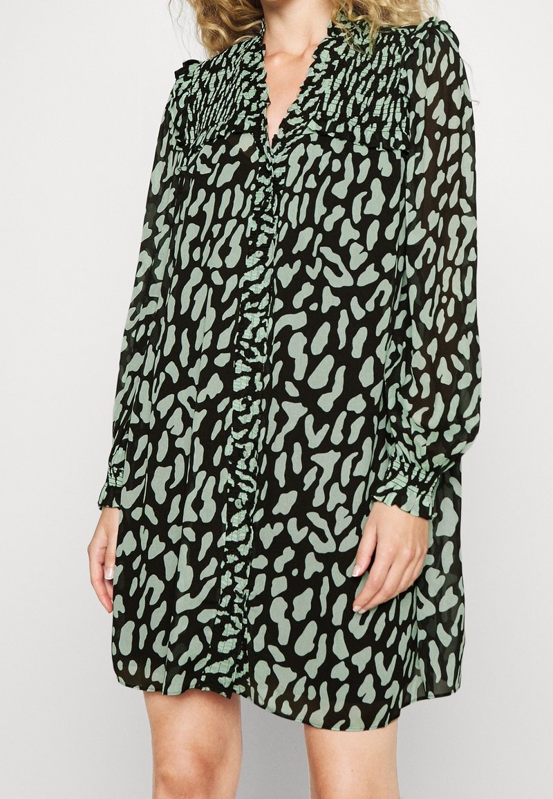 Vihreä ja musta nappiversio mekosta, jossa on kuviollinen suunnittelu, röyhelökaulus ja pitkät hihat, joissa on elastiset hihansuut. Kevyt kangas.